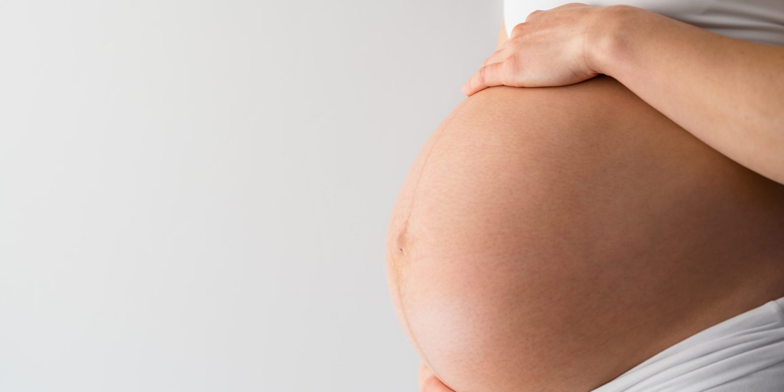 Midsection of unrecognizable standing mother gently holding her very round pregnant baby belly. Side angle view. White background. Bright shot. salud bucal durante el embarazo